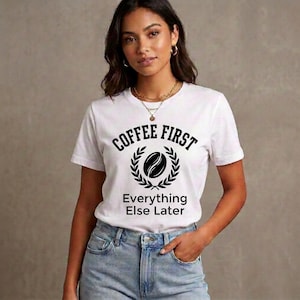 May include: A white short-sleeved t-shirt featuring a black graphic print. The design includes the text COFFEE FIRST at the top, a central coffee bean icon surrounded by a laurel wreath, and the text Everything Else Later at the bottom.