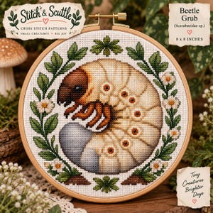 Puede incluir: Un bastidor de bordado circular de madera que contiene un patrón de punto de cruz de una larva de escarabajo de color crema con cabeza y patas de color marrón oscuro, rodeada por una corona de hojas verdes y pequeñas flores blancas.