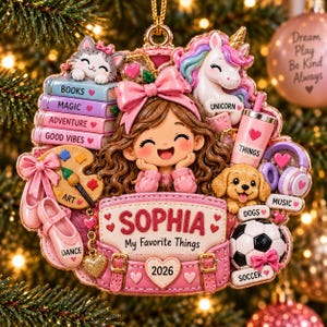 May include: A decorative ornament featuring a central figure with brown hair and a pink bow, surrounded by icons including a cat, unicorn, books labeled Books, Magic, Adventure, Good Vibes, an art palette, ballet shoes, a dog, headphones, and a soccer ball. The center displays the name Sophia, My Favorite Things, and the year 2026.