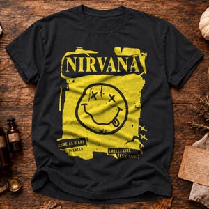Può includere: Una t-shirt nera a maniche corte con una stampa grafica gialla. Il design include la parola NIRVANA in alto, una grande faccina sorridente con occhi a forma di X e il testo COME AS U ARE THEATER e SMELLS LIKE TEEN SPIRIT in basso.