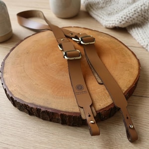 May include: A tan leather camera strap featuring adjustable brass-toned metal buckles and hardware. The strap includes a debossed circular logo with the text noryshan and the phrase made in new hampshire printed on the surface of the leather.