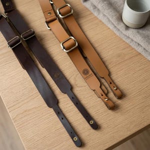 May include: Two leather camera straps, one dark brown and one tan, featuring adjustable metal buckles and rivets. Each strap is embossed with the circular logo and text 'holydust'. The straps have reinforced stitching and pre-punched holes at the ends.