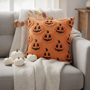Peut inclure: Un coussin carré orange orné d'un motif répétitif de visages de citrouilles d'Halloween noirs en relief. Le coussin est présenté avec trois petites citrouilles décoratives blanches et un plaid en tricot clair avec une branche de feuilles.