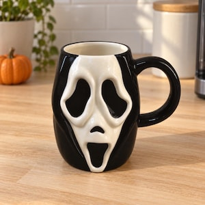 Peut inclure: Une tasse en céramique présentant un design moulé en trois dimensions du masque Ghostface de la franchise de films d'horreur Scream. La tasse est principalement noire avec un visage sculpté en relief blanc montrant des yeux creux.