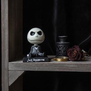 Peut inclure: Une figurine de Jack Skellington avec une grande tête de crâne blanc, des yeux noirs creux et une bouche cousue. Le personnage porte un costume à rayures noires et blanches et est assis sur une base rectangulaire noire avec le texte Jack Skellington.