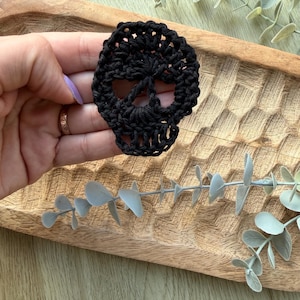 May include: A black crochet skull applique featuring an openwork design for the eye sockets and jaw. The piece is crafted from dark yarn with visible stitching, forming a distinct skull shape with a rounded cranium and a defined lower jaw area.