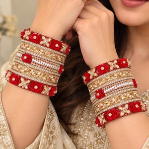 Peut inclure: Un ensemble de bracelets joncs superposés ornés de fleurs rouges éclatantes, de cristaux transparents et de bandes en métal doré. Les bracelets sont disposés en une large structure multicouche alternant rangées de pierres et fleurs.