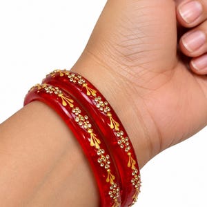Puede incluir: Dos brazaletes rojos translúcidos con un patrón floral dorado repetitivo acentuado con pequeños cristales transparentes. Las pulseras circulares tienen un acabado liso y pulido y se usan juntas en la muñeca, mostrando el color rojo vibrante.