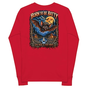 May include: A red long-sleeve shirt featuring a graphic on the back of a bat wearing a cap, flying over a lake and cabins at night under a full moon. The text reads BORN TO BE BATTY at the top and STAY WILD STAY MISFIT at the bottom.
