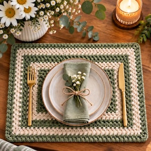 May include: A rectangular crochet placemat featuring alternating bands of sage green and cream yarn. Centered on the placemat is a speckled ceramic plate topped with a folded sage green linen napkin, a small sprig of greenery, and a gold fork and knife.
