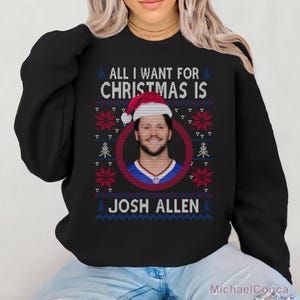 Puede incluir: Una sudadera negra de manga larga con un estampado gráfico que incluye el texto ALL I WANT FOR CHRISTMAS IS JOSH ALLEN. El diseño presenta un retrato circular de Josh Allen con un gorro de Papá Noel, rodeado de patrones de copos rojos y blancos.