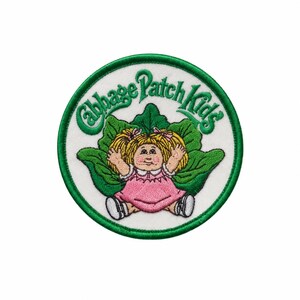 May include: A circular embroidered patch featuring the text Cabbage Patch Kids in green thread above a cartoon character with blonde pigtails, wearing a pink dress, sitting in front of large green leaves, all on a white background with a green border.