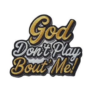 Może przedstawiać: Haftowana naszywka z napisem God Don't Play Bout' Me! wykonanym stylizowaną czcionką. Słowo God oraz fraza Bout' Me! są wyszyte metaliczną złotą nicią, natomiast Don't Play białą nicią, całość wykończona czarnym obramowaniem.