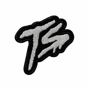 Puede incluir: Un parche bordado que presenta las letras TS en una fuente estilizada inspirada en el grafiti. Las letras están cosidas con hilo blanco sobre un fondo negro sólido, con un borde negro grueso que delinea la forma del diseño.