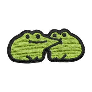May include: An embroidered patch featuring two lime green frogs with black outlines. The frogs are positioned side-by-side, with the left frog appearing to lean toward the right frog. The design is rendered in a textured stitch pattern.