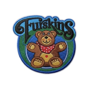 Puede incluir: Un parche bordado que muestra un oso de peluche marrón con un pañuelo rojo de lunares blancos. El oso está centrado sobre un fondo circular azul y verde. La palabra "Fruiskins" está bordada en letras cursivas negras sobre el oso.