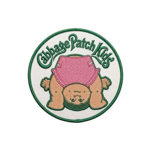 May include: A circular embroidered patch featuring the green text Cabbage Patch Kids arched over an illustration of a baby with tan skin, dark hair, and pink ruffled diaper. The design is set against a white background with a green border.