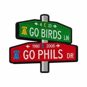 Op de afbeelding: Een geborduurde patch met twee straatnaamborden op een grijze paal. Het bovenste groene bord toont GO BIRDS LN met een gele bel en 4 E 33. Het onderste rode bord toont GO PHILS DR met een blauwe bel en 1980 E 2008. Beide hebben zwarte randen.