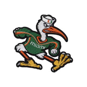 May include: An embroidered patch featuring a white ibis mascot wearing a white sailor hat with an orange U logo. The ibis wears a green jersey with orange trim and the word Miami in white text, featuring orange legs and yellow feet.