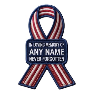 May include: A ribbon-shaped embroidered patch featuring red and white vertical stripes with a dark blue border. A central dark blue rectangular section contains the white embroidered text: IN LOVING MEMORY OF ANY NAME NEVER FORGOTTEN.