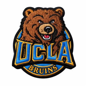 May include: An embroidered patch featuring a brown bear head with a black nose and open mouth, positioned above the blue and gold text UCLA and BRUINS. The design is set against a circular blue background with a thick black border outline.