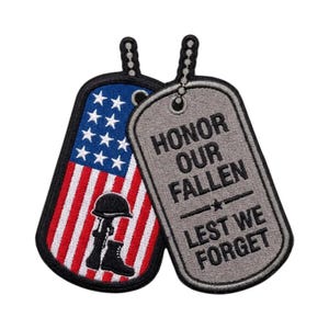Peut inclure: Deux écussons brodés en forme de plaques d'identité militaires. L'un représente le drapeau américain avec une silhouette noire de casque et de fusil. L'autre, gris, porte le texte noir HONOR OUR FALLEN LEST WE FORGET avec une étoile.