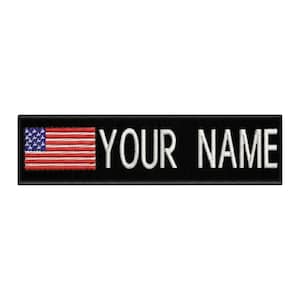 May include: A rectangular black embroidered patch featuring a small American flag on the left side with red, white, and blue thread. To the right of the flag, the text YOUR NAME is stitched in bold, white, capital sans-serif lettering.
