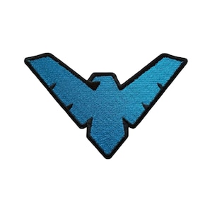 Op de afbeelding: Een blauwe geborduurde patch in de vorm van een gestileerde vogel of chevron-symbool. Het ontwerp heeft een felblauw midden met een donkerder blauw verloop-effect, omlijnd door een dikke, solide zwarte rand met een textielachtige afwerking.