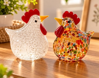 May include: Two glass chicken figurines. One is clear with white speckles, and the other features a multicolored confetti pattern. Both have bright red combs and wattles, yellow beaks, and small black eyes, standing as decorative glass sculptures.