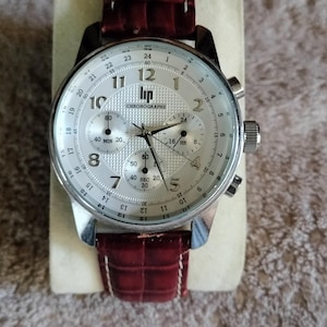 Puede incluir: Un reloj cronógrafo de tono plateado con esfera blanca texturizada, que presenta el logotipo de Lip y la palabra Chronographe. Tiene números arábigos negros, tres subesferas y una correa de cuero rojo oscuro con patrón de cocodrilo y costuras blancas.