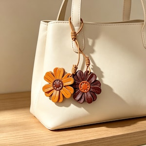 May include: A cream-colored handbag featuring two dangling flower-shaped charms attached to the handle. One charm is bright orange with a dark brown center, and the other is dark brown with an orange center, both connected by thin leather cords.
