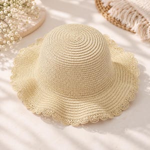 May include: A light beige straw hat featuring a rounded crown and a wide, flexible brim. The edge of the brim is finished with a delicate, scalloped crochet lace trim in a matching beige tone, creating a circular, textured pattern throughout.