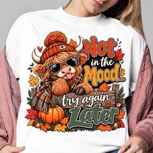Peut inclure: Un t-shirt blanc à manches longues avec un motif de vache Highland portant un bonnet orange brûlé, des lunettes à motif léopard et une écharpe à carreaux. La vache tire la langue. Texte : Not in the Mood... try again Later, avec des citrouilles.