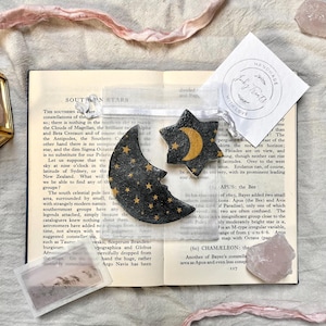 May include: A black crescent moon and a black star, both decorated with gold celestial patterns, are inside a sheer white organza pouch. The pouch rests on an open book page titled Southern Stars, accompanied by a small white tag with text.