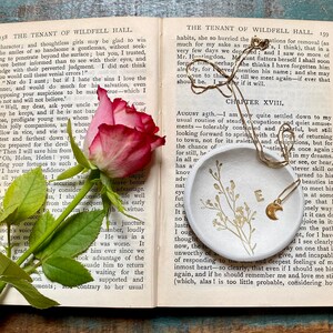 May include: A white ceramic dish with a gold crescent moon charm necklace on top. The dish has a floral design and the necklace has a gold letter 'E' charm. The dish is sitting on a book with the text 'The Tenant of Wildfell Hall' visible.