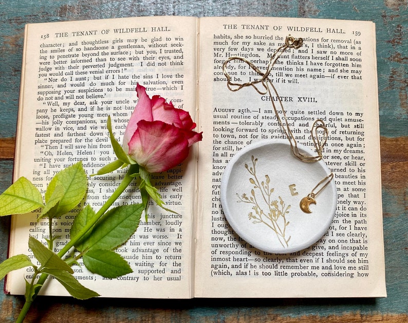 May include: A white ceramic dish with a gold crescent moon charm necklace on top. The dish has a floral design and the necklace has a gold letter 'E' charm. The dish is sitting on a book with the text 'The Tenant of Wildfell Hall' visible.