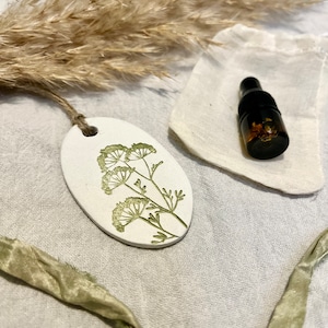 May include: An off-white ceramic tag with a green floral print, a small brown glass bottle, and a small white pouch. The tag is attached to a brown string. Dried grass and a green ribbon are also visible.