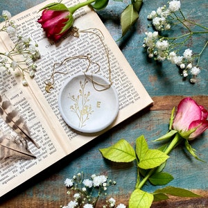 May include: A white ceramic ring dish with a gold floral design and the letter "E" in the center. The dish is sitting on an open book with a rose and white flowers in the background.