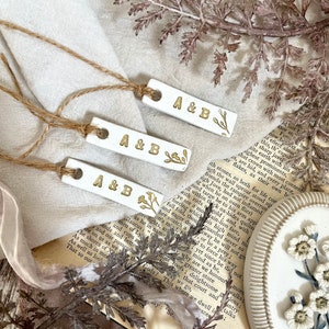 May include: Three white ceramic gift tags with gold lettering and floral designs. Each tag reads "A&B". The tags are tied with twine.