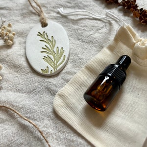 May include: An oval, white ceramic essential oil diffuser with a green botanical design, suspended by a twine string. A small amber glass bottle with a black dropper top sits next to a small, off-white drawstring bag.