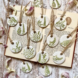 May include: A set of 10 ceramic ornaments featuring different types of green foliage, each with a hole for hanging. The ornaments are white with green leaf designs and are displayed on a book with text about cuckoos.