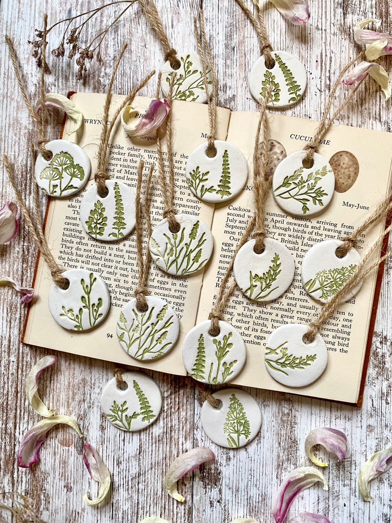 May include: A set of 10 ceramic ornaments featuring different types of green foliage, each with a hole for hanging. The ornaments are white with green leaf designs and are displayed on a book with text about cuckoos.