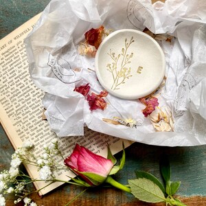 May include: A white ceramic ring dish with a gold floral design and the letter "E" in the center. The dish is surrounded by white tissue paper and dried flower petals.