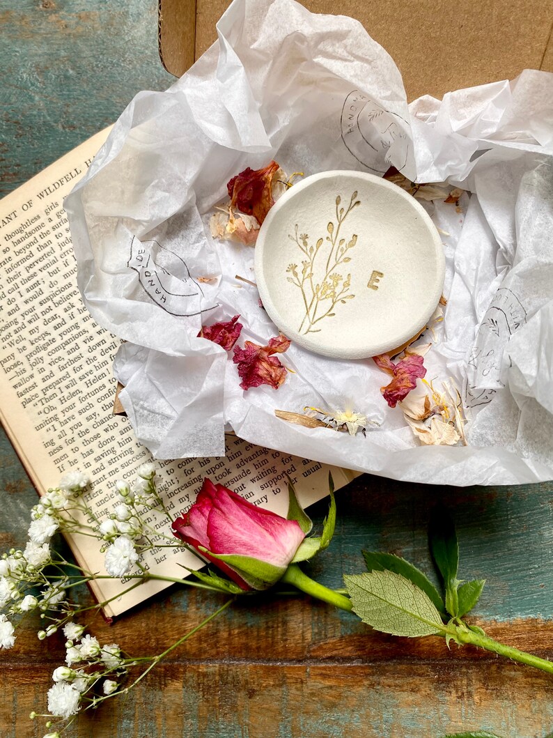 May include: A white ceramic ring dish with a gold floral design and the letter "E" in the center. The dish is surrounded by white tissue paper and dried flower petals.