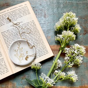 May include: A white ceramic dish with a gold chain and a crescent moon charm. The dish has a floral design and the letter "E" in gold. The dish is sitting on an open book with white flowers on a wooden surface.