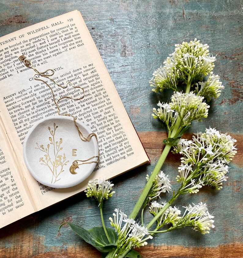 May include: A white ceramic dish with a gold chain and a crescent moon charm. The dish has a floral design and the letter "E" in gold. The dish is sitting on an open book with white flowers on a wooden surface.