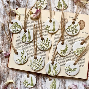 May include: A set of ten white ceramic ornaments with green botanical illustrations. Each ornament is round and has a hole at the top for hanging. The ornaments are arranged on a wooden surface with a book open to a page about cuckoos.
