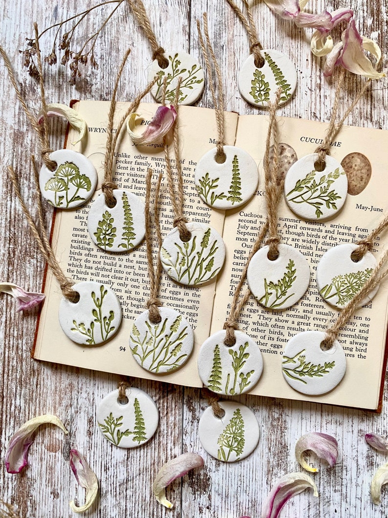 May include: A set of ten white ceramic ornaments with green botanical illustrations. Each ornament is round and has a hole at the top for hanging. The ornaments are arranged on a wooden surface with a book open to a page about cuckoos.