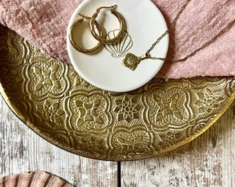 May include: A white ceramic trinket dish with a seashell design, holding gold hoop earrings and a gold chain necklace with a pendant. The dish is sitting on a gold tray with an ornate design.