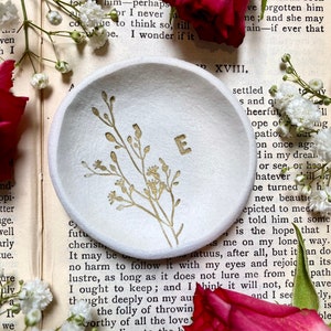 May include: A white ceramic ring dish with a gold floral design and the letter "E" in the center. The dish is sitting on a page of a book with text.